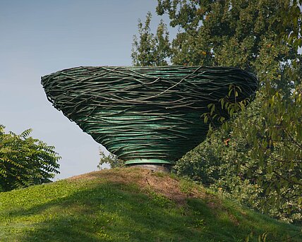 Kienzers Skulptur aus gewundenem Kupferrohr ragt über der Hügelkuppe auf wie ein riesiges Vogelnest. Das Objekt wirkt dadurch wie eine spiegelbildliche oder umgekehrte Fortsetzung des Hügels. Das Material bezieht sich außerdem auf den ewigen Kreislauf der Veränderung in der Natur, indem es sich durch Oxidation selbst verändert und ein Teil der Natur wird.