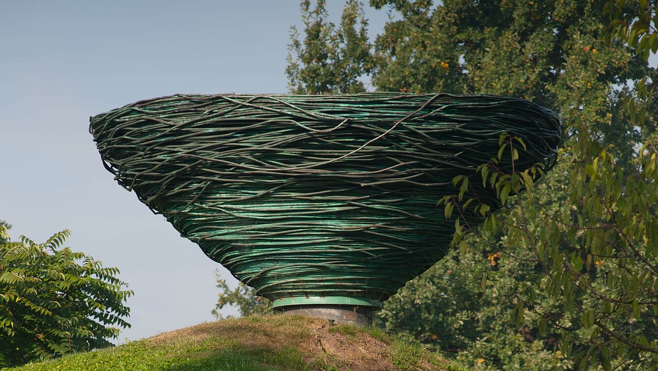 Kienzers Skulptur aus gewundenem Kupferrohr ragt über der Hügelkuppe auf wie ein riesiges Vogelnest. Das Objekt wirkt dadurch wie eine spiegelbildliche oder umgekehrte Fortsetzung des Hügels. Das Material bezieht sich außerdem auf den ewigen Kreislauf der Veränderung in der Natur, indem es sich durch Oxidation selbst verändert und ein Teil der Natur wird.