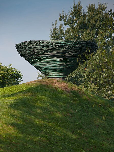 Kienzers Skulptur aus gewundenem Kupferrohr ragt über der Hügelkuppe auf wie ein riesiges Vogelnest. Das Objekt wirkt dadurch wie eine spiegelbildliche oder umgekehrte Fortsetzung des Hügels. Das Material bezieht sich außerdem auf den ewigen Kreislauf der Veränderung in der Natur, indem es sich durch Oxidation selbst verändert und ein Teil der Natur wird.