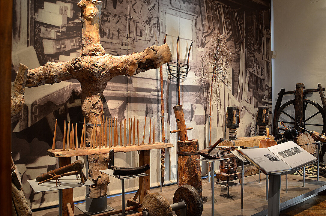 Photograph of an exhibition space. On a pedestal lie and stand various objects made of wood. In the background there is a photo of a workshop.
