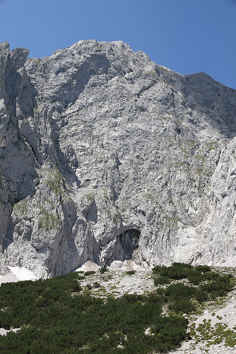 Foto von einer Felswand mit grüner Wiese und blauem Himmel. In der Felswand befindet sich ein torförmige Grube.