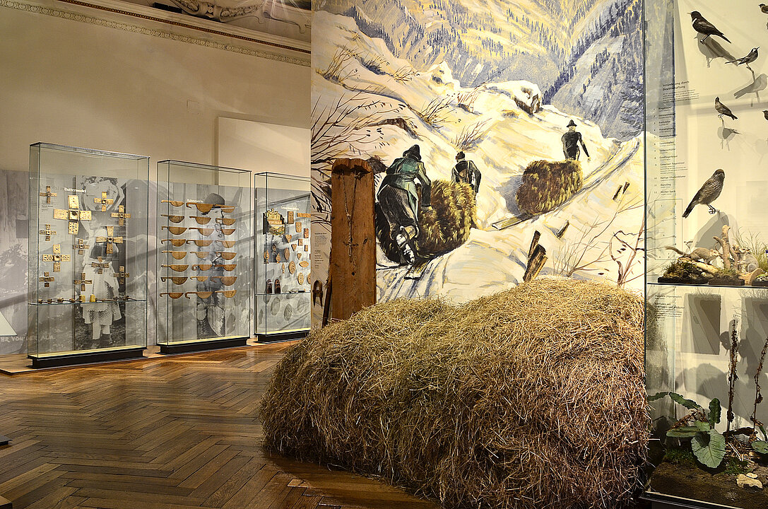 Photography of an exhibition space. In front of a painted wall is a large pile of hay. On the painted wall are depicted people in a winter landscape. These bring hay on sledges into the valley.