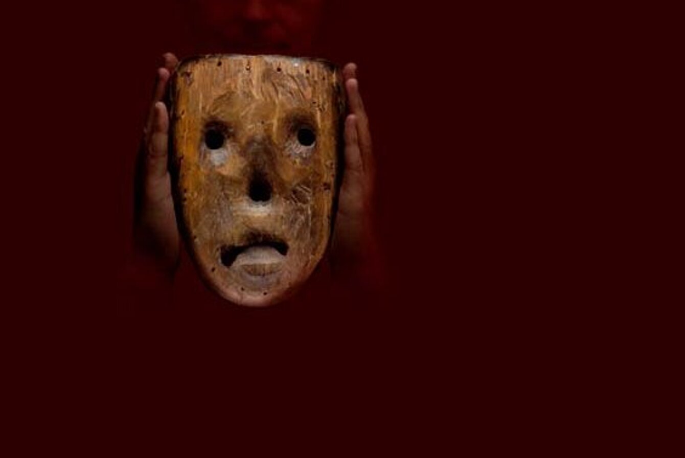 Das Foto ist fast komplett schwarz. Nur zwei Hände sind zu erkennen, eine Maske aus Holz halten. Von der Maske ist nur die Innenseite zusehen. Mit Löchern für einen Mund, zwei Augen und die Nase.