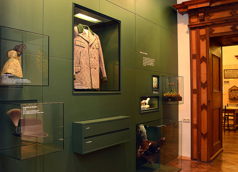 Foto von einem Ausstellungsraum. In eine grüne Wand sind Vitrinen eingelassen. In ihnen ist ein Mantel (Schladminger Rock), eine Goldhaube und Tiere z.B. ein Wiesel zu sehen.