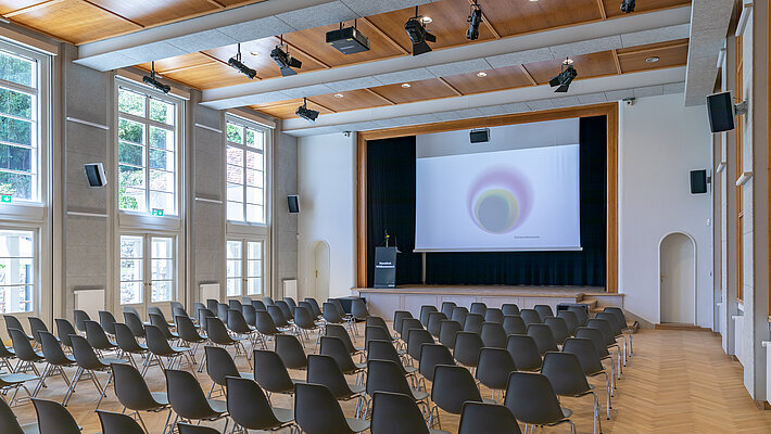 Sessel stehen in Richtung Bühne im Heimatsaal, auf dieser ist ein Rednerpult sowie eine Leinwand.