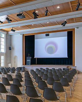 Chairs stand in the direction of the stage in the Heimatsaal, on it is a lectern as well as a screen.