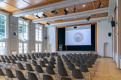 Chairs stand in the direction of the stage in the Heimatsaal, on it is a lectern as well as a screen.