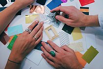 Auf dem Bild im Querformat sind vier Hände zu sehen, welche auf unterschiedlichem Material wie Holz oder Papier ruhen. 