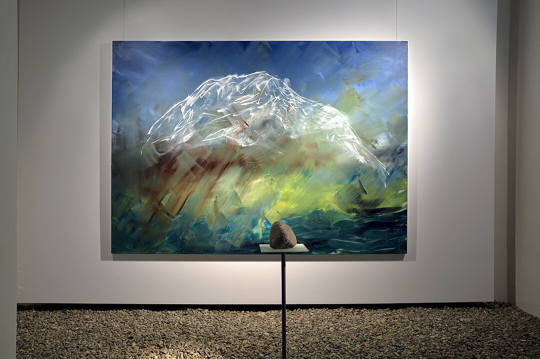 Photo of an exhibition view. In the middle of the room is a plinth with a limestone. Behind it hangs a painting showing Mount Grimming in abstract style.