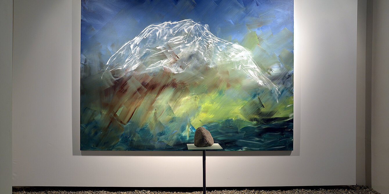 Photo of an exhibition view. In the middle of the room is a plinth with a limestone. Behind it hangs a painting showing Mount Grimming in abstract style.