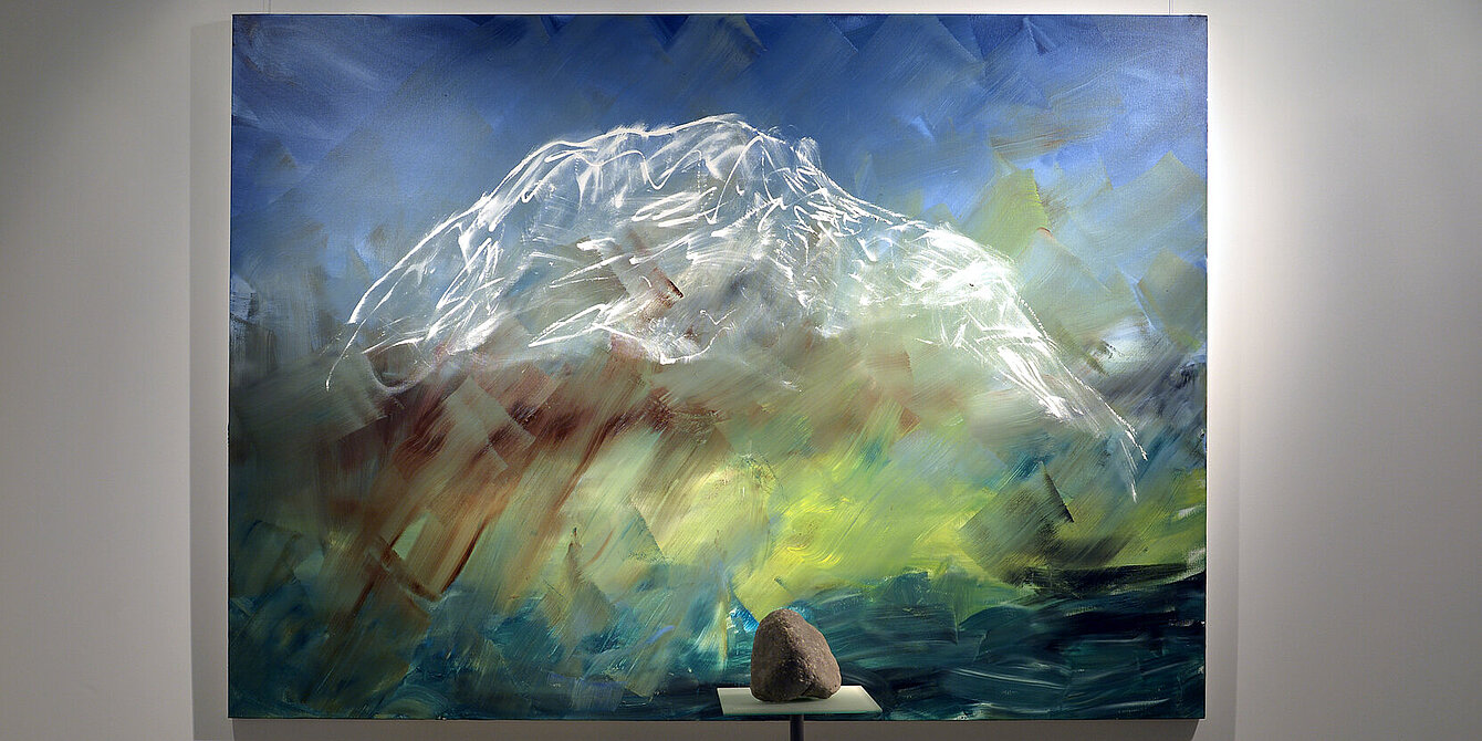 A limestone rock lies on a pedestal in front of a painting. The painting shows the mountain Grimming.