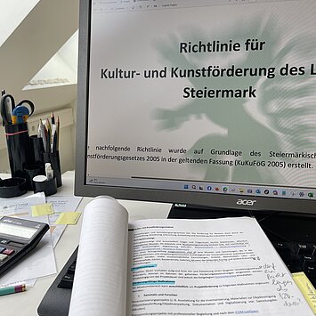 Blick auf Schreibtisch mit Monitor, auf dem die neue Kunst- und Kulturförderrichtlinie des Landes Steiermark zu sehen ist.