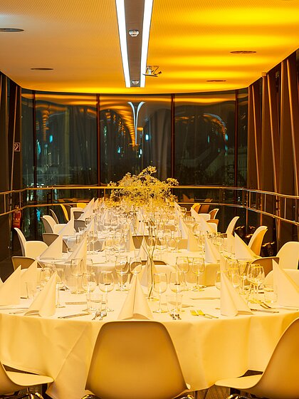 In der Needle im Kunsthaus Graz sind runde Galatische eingedeckt. Der Raum ist in goldenes Licht getaucht, auf den Tischen steht passende Tischdekoration.