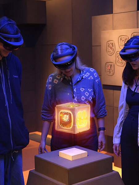 Eine Gruppe jugendlicher Besucher:innen mit AR-Brillen betrachtet eine virtuelle, leuchtende Box im AR-Raum des CoSAs.