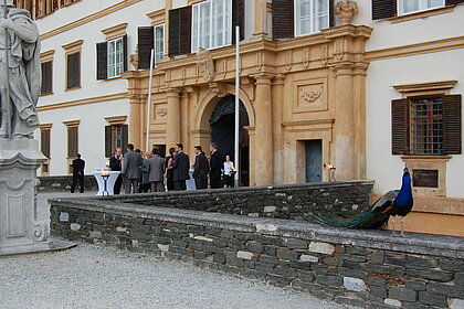 Menschen stehen am Vorplatz des Schloss Eggenberg bei einem Empfang. Ein Pfau sitzt daneben auf einer Mauer.
