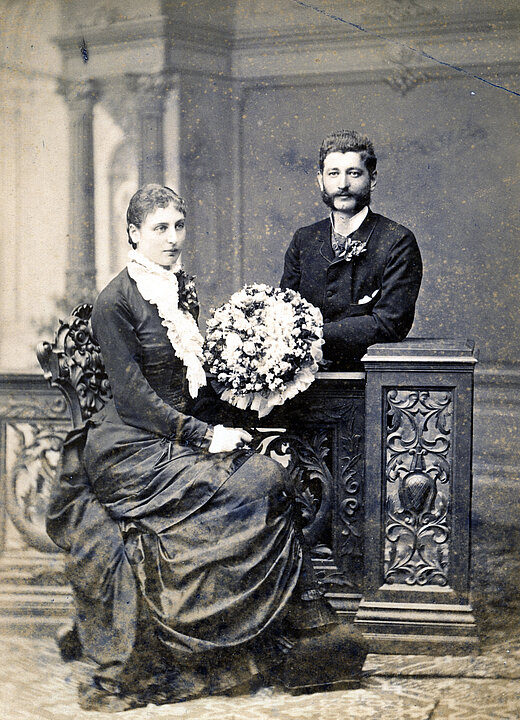
An old wedding photo. The bride wears a dark dress and holds a large bouquet of flowers. The man is standing behind there and is wearing a dark suit. They both smile very slightly.