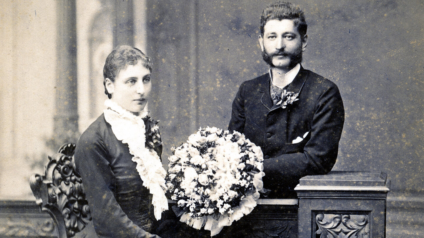 Ein altes Hochzeitfoto. Die Braut trägt ein dunkles Kleid und hält einen großen Blumenstrauß. Der Man steht da hinter und trägt einen dunklen Anzug. Beide lächeln ganz leicht.