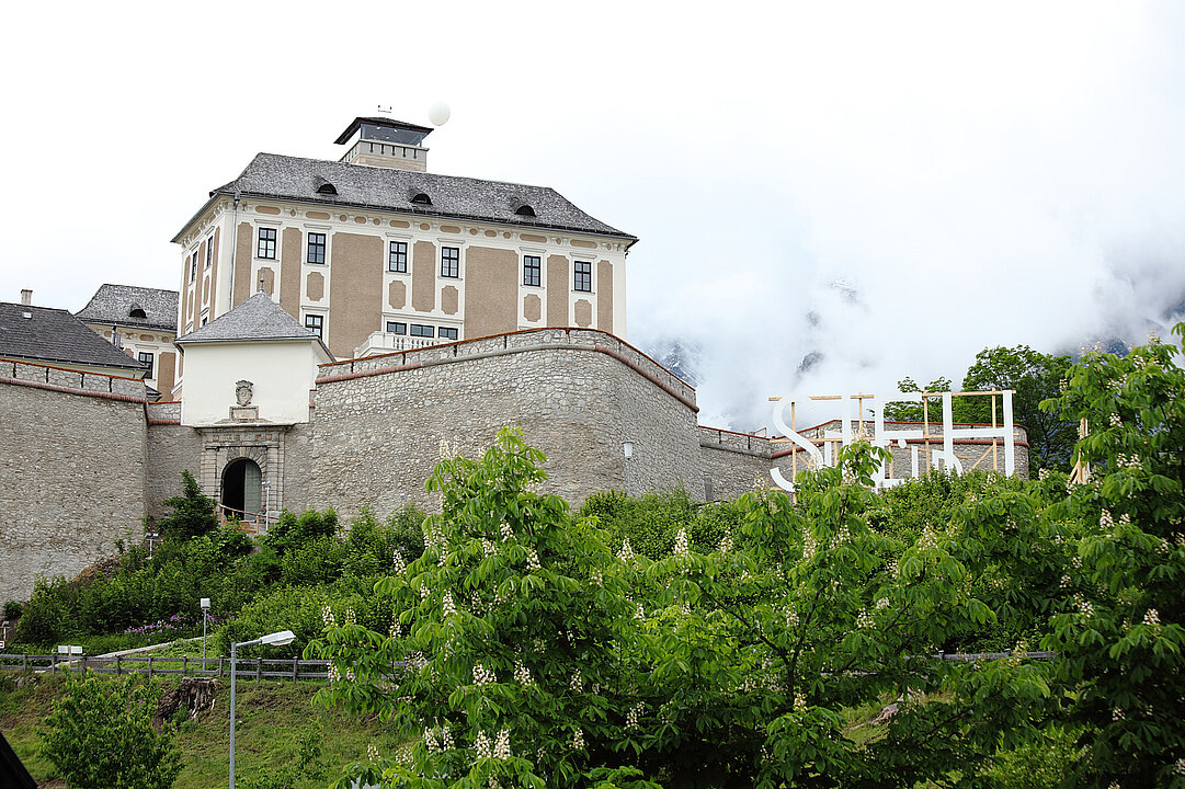 Fotografie von Schloss Trautenfels. Am Fuße des Schlossbergs blühen Kastanien, unterhalb der Schlossmauer steht eine große Installation aus weiß gestrichenem Holz, das den Schriftzug "Sieh" ergibt.