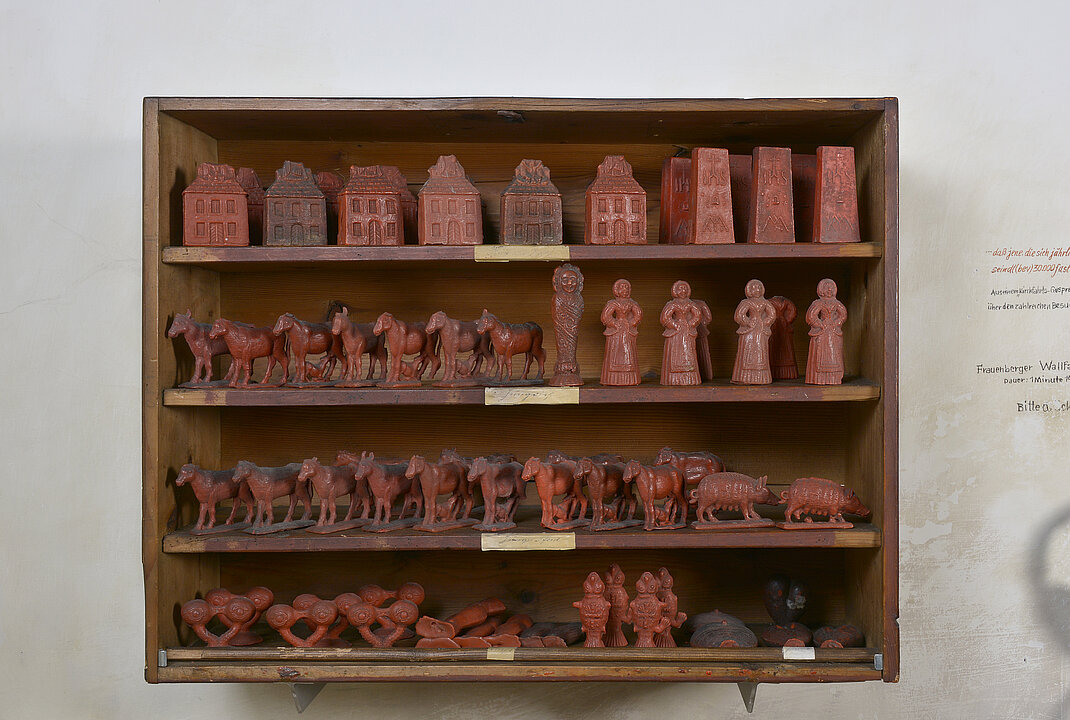 Wooden box in which there are numerous lined up wax figurines showing horses, eyes, people and buildings.