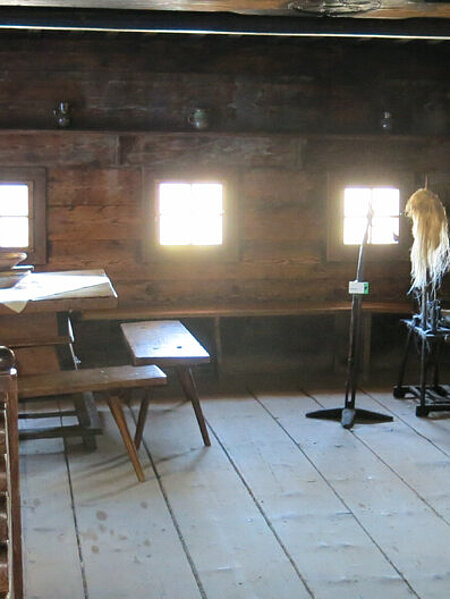 The large parlor with bed, farmhouse table, pinewood holder and cradle. The small windows are brightly lit by the sun shining in from outside.