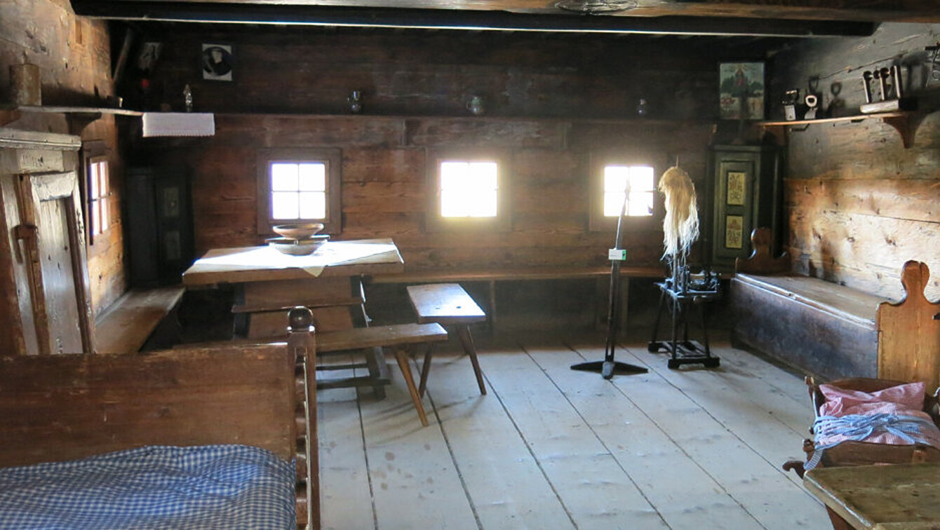 The large parlor with bed, farmhouse table, pinewood holder and cradle. The small windows are brightly lit by the sun shining in from outside.