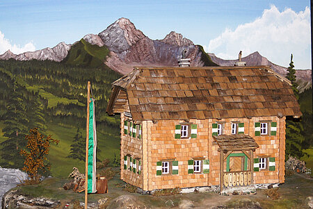 Foto von einem Holzmodell von einer Hütte, die vor einer gezeichneten Berglandschaft steht.