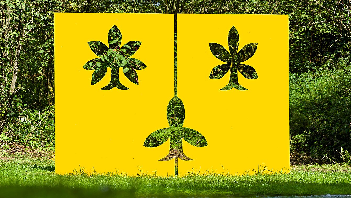 The sculpture is a vertical yellow metal plate with three leaf-shaped cut-outs.