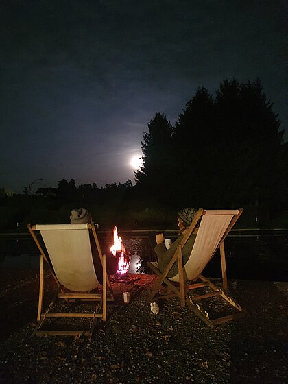 Two people sit at night in the sculpture park in front of a fire barrel. In the background the full moon rises.