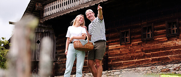 Ein Mann und eine Frau mit Picknickskorb vor dem Geburtshaus stehend. Der Mann zeigt mit erhobener Hand in die Ferne
