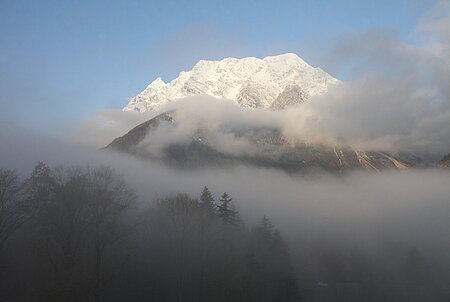 Foto von dem Berg Grimming. Die Schnee bedeckte Spitze ragt aus den Wolken und dem Nebel hervor und wird von der Sonne beleuchtet.