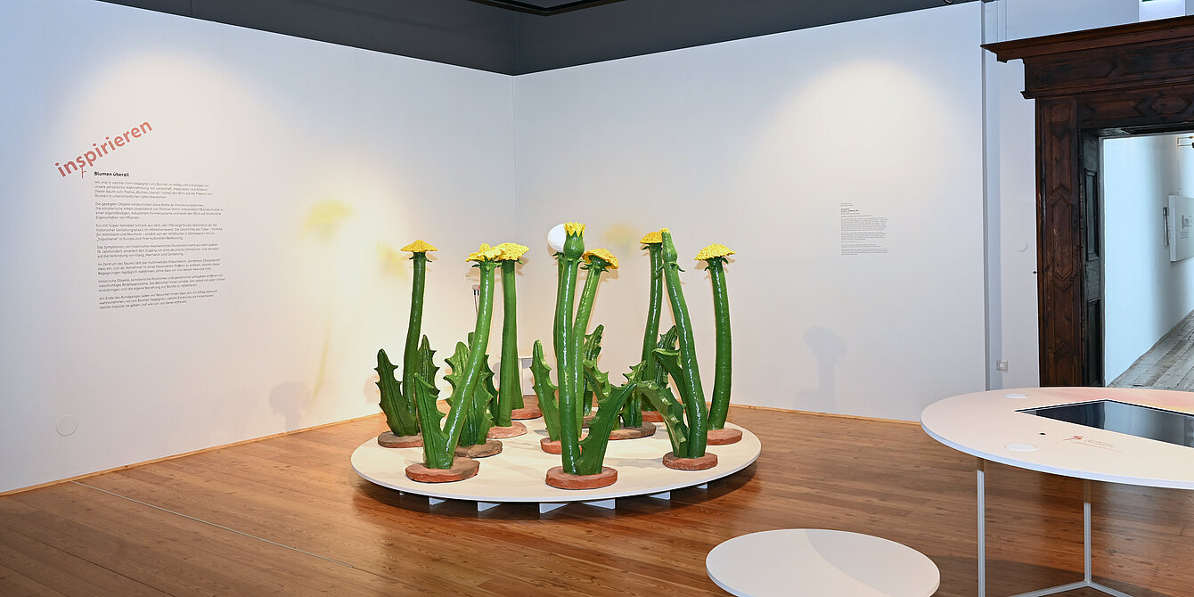 Ausstellungsraum mit hellem Holzboden und weißen Wänden. In der Mitte steht auf einer runden weißen Plattform eine Installation aus mehreren übergroßen, grünen Löwenzahnpflanzen. Links befindet sich eine Wandbeschriftung mit dem Titel „inspirieren“ und erläuterndem Text. Rechts sind ein interaktiver Tisch mit Bildschirm sowie ein weißer Hocker zu sehen.