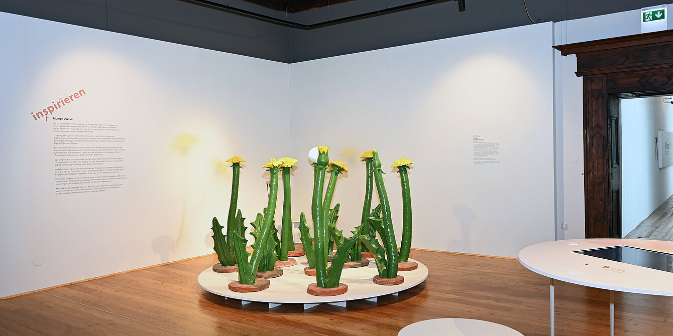 Ausstellungsraum mit hellem Holzboden und weißen Wänden. In der Mitte steht auf einer runden weißen Plattform eine Installation aus mehreren übergroßen, grünen Löwenzahnpflanzen. Links befindet sich eine Wandbeschriftung mit dem Titel „inspirieren“ und erläuterndem Text. Rechts sind ein interaktiver Tisch mit Bildschirm sowie ein weißer Hocker zu sehen.