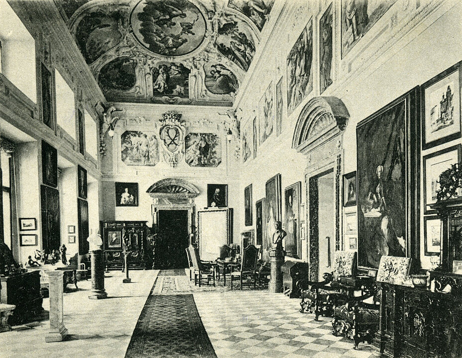 
Old photograph of the Marble Hall. Numerous paintings hang on the walls. There is a long carpet in the room and there is furniture along the walls.