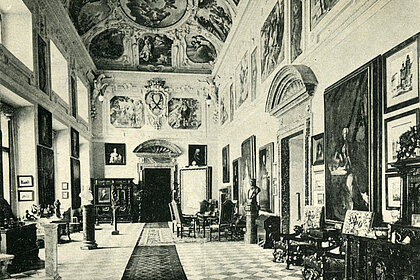 
Old photograph of the Marble Hall. Numerous paintings hang on the walls. There is a long carpet in the room and there is furniture along the walls.