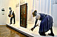 Photo of an exhibition room. On a long pedestal is a door. This is painted with motifs from the four seasons. To the right and left of the door are two mannequins in peasant clothes, working.