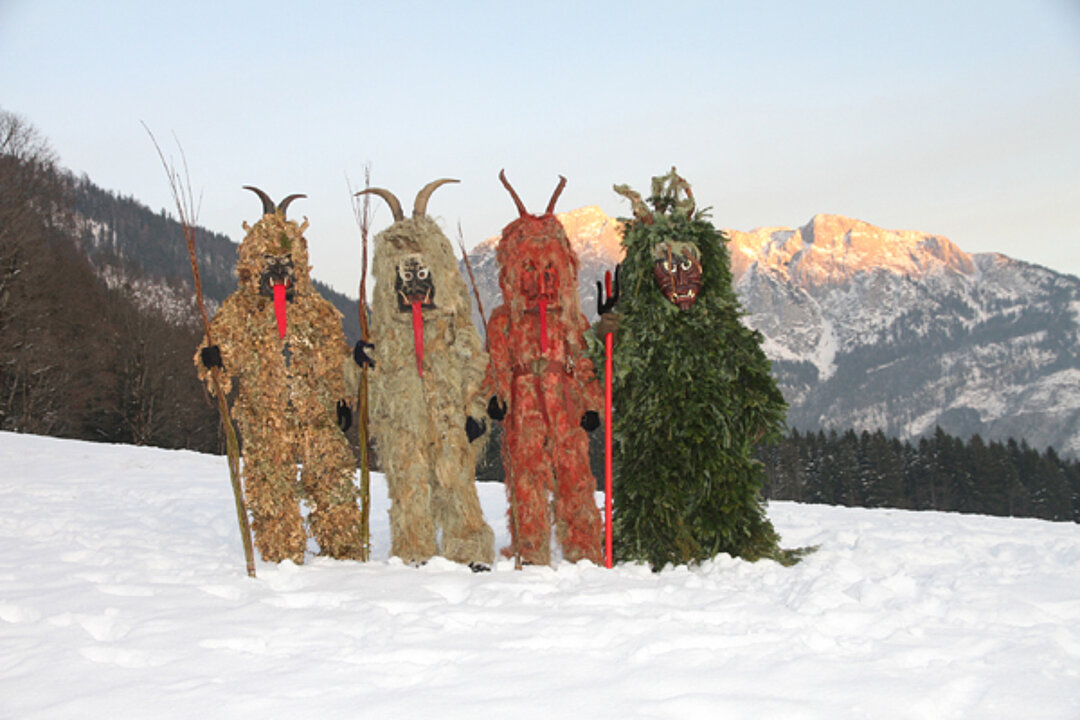 Vier verkleidete Figuren stehen auf dem Schnee. Im Hintergrund ist ein Berg zu sehen. Die Figuren haben Hörner und lange Rote Zungen aus Stoff, die ihnen aus den Mündern hängen.