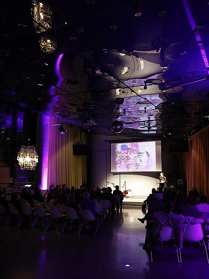 Der Space04 im Kunsthaus Graz ist farblich beleuchtet. Es sitzen Menschen im Publikum, auf dem Podium wird eine Ansprache gehalten.