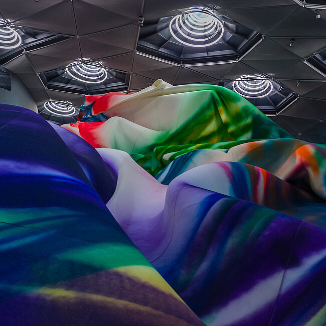 Das Bild zeigt eine Szene aus dem Space01 des Kunsthauses Graz, in dem Werke der Künstlerin Katharina Grosse ausgestellt sind. Farben werden unscharf über Leinwände, Landschaften und funktionale Objekte gelegt, wodurch die Grenzen zwischen ihnen verschwimmen. Böden scheinen organisch in das Bild zu wachsen, während Räume und Objekte eine skulpturale Erscheinung annehmen.