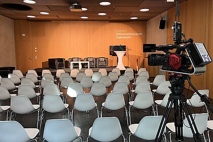 Eine Reihenbestuhlung ist im Auditorium vor dem Podium aufgebaut. Auf diesem befinden sich Couches für die Gesprächsgäste. Im Vordergrund steht eine Kamera für Filmaufnahmen.