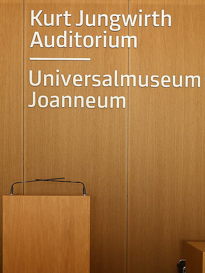Das Foto zeigt die hölzerne Rückwand im Kurt Jungwirth Auditorium im Joanneumsviertel. Im Vordergrund ist das Rednerpult im Hintergrund sieht man den Schriftzug "Kurt Jungwirth Auditorium, Universalmuseum Joanneum".