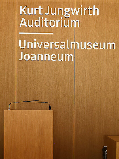 The photo shows the wooden back wall in the Kurt Jungwirth Auditorium in the Joanneumsviertel. In the foreground is the speaker´s podium; in the background, you can see the inscription “Kurt Jungwirth Auditorium, Universalmuseum Joanneum.”