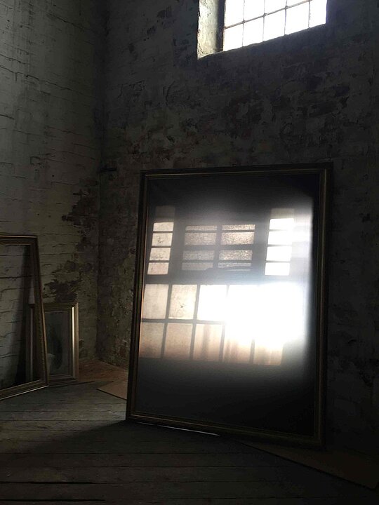 In einem dunklen Raum steht ein Spiegel, der ein Fenster und das dadurch einfallende Licht einfängt.