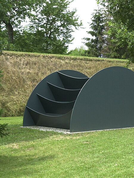 The sculpture consists of a large, dark gray, semi-circular figure and has pockets or compartments with a slightly sagging base. These are attached to a kind of wheel that faces the viewer like an uncomfortable ladder. The reference to the body is derived from the perception of the object as an obstacle.