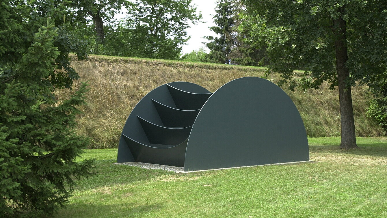 The sculpture consists of a large, dark gray, semi-circular figure and has pockets or compartments with a slightly sagging base. These are attached to a kind of wheel that faces the viewer like an uncomfortable ladder. The reference to the body is derived from the perception of the object as an obstacle.