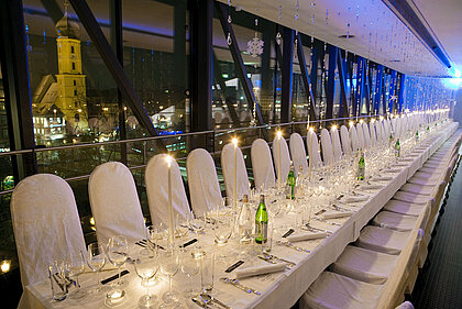 In den Räumlichkeiten der Needle im Kunsthaus Graz erstreckt sich eine lange Gala-Tafel, gedeckt für ein Abendessen mit Kerzen und Ambientelicht. Im Hintergrund sieht man die Stadt Graz bei Nacht.