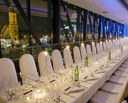 In den Räumlichkeiten der Needle im Kunsthaus Graz erstreckt sich eine lange Gala-Tafel, gedeckt für ein Abendessen mit Kerzen und Ambientelicht. Im Hintergrund sieht man die Stadt Graz bei Nacht.