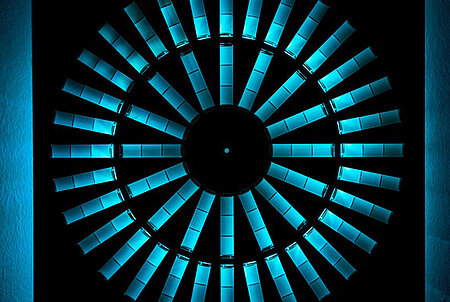 Foto von einer Kunstinstallation: Aus einer schwarzen Platte sind kreisförmig Rechtecke ausgeschnitten. Dahinter ist ein blaues Licht, dass durch die Aussparrungen fällt. 