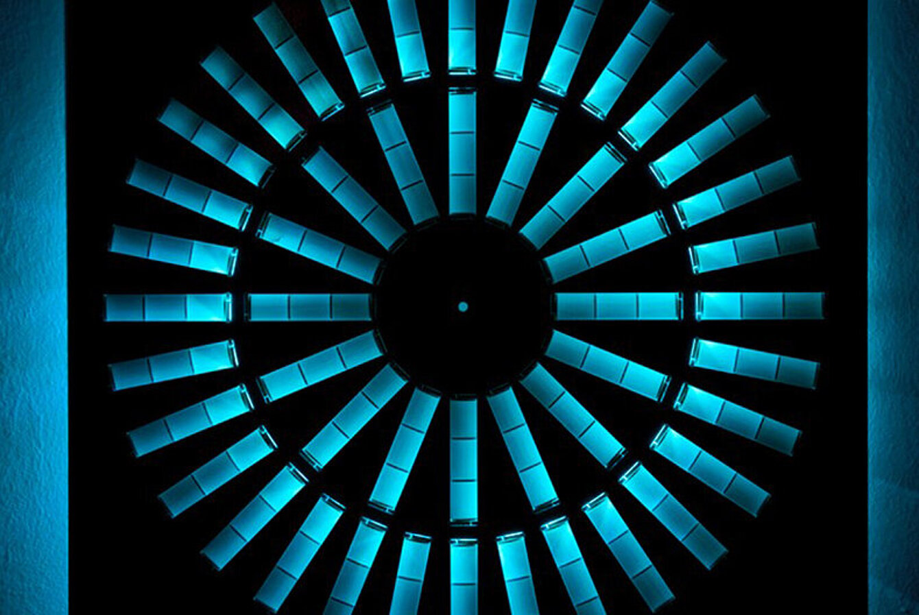 Foto von einer Kunstinstallation: Aus einer schwarzen Platte sind kreisförmig Rechtecke ausgeschnitten. Dahinter ist ein blaues Licht, dass durch die Aussparrungen fällt. 
