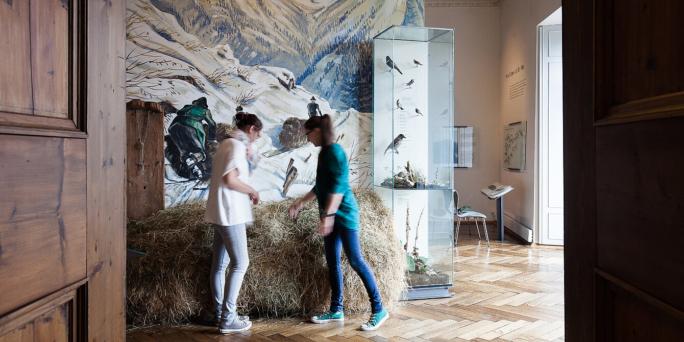 two women stand in a room in front of a haystack and look at it