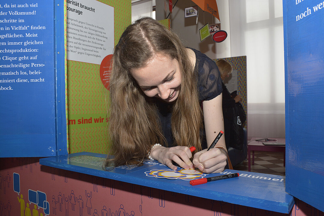 Eine Frau steht in einem inszenierten Ausstellungsraum und schreibt etwas auf eine Karte.